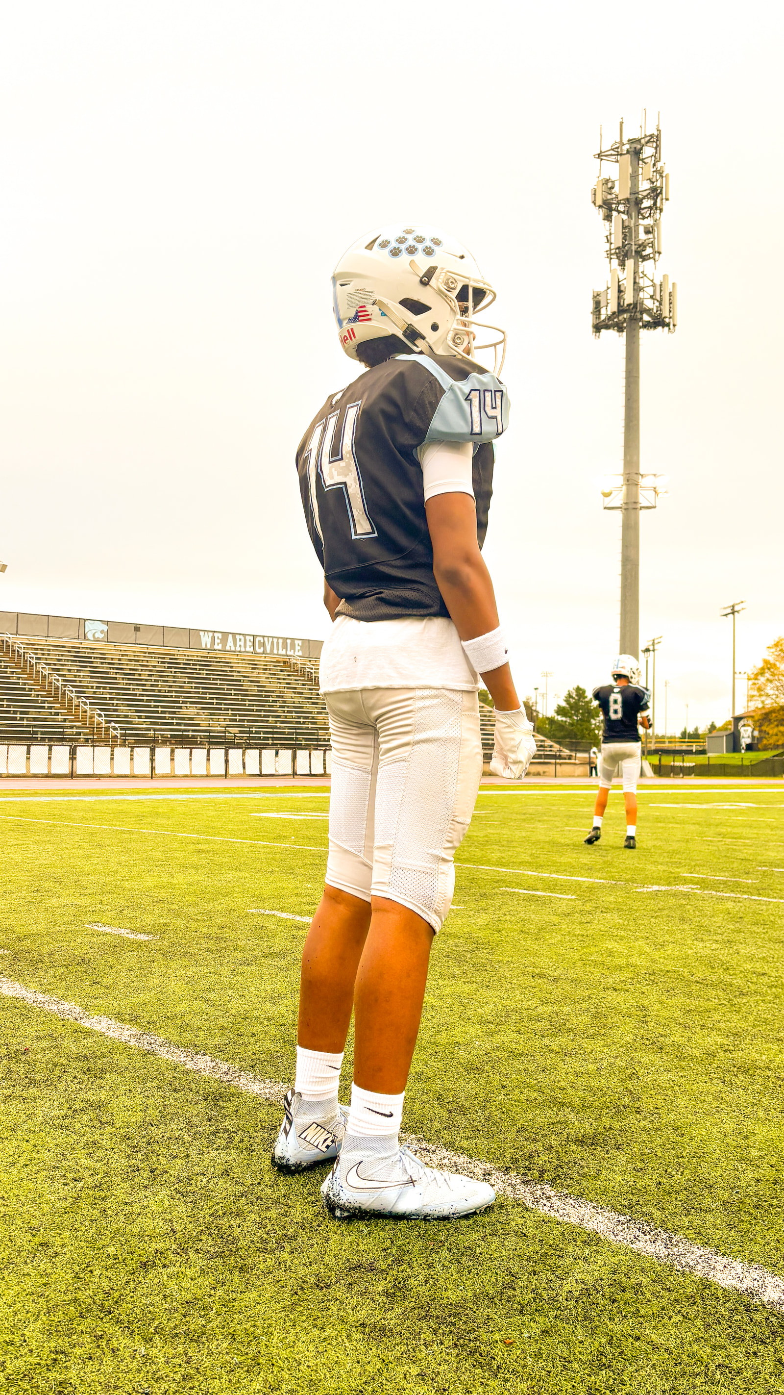 A football player stands on a field facing away from the camera, with a goalpost and empty bleachers in the background.