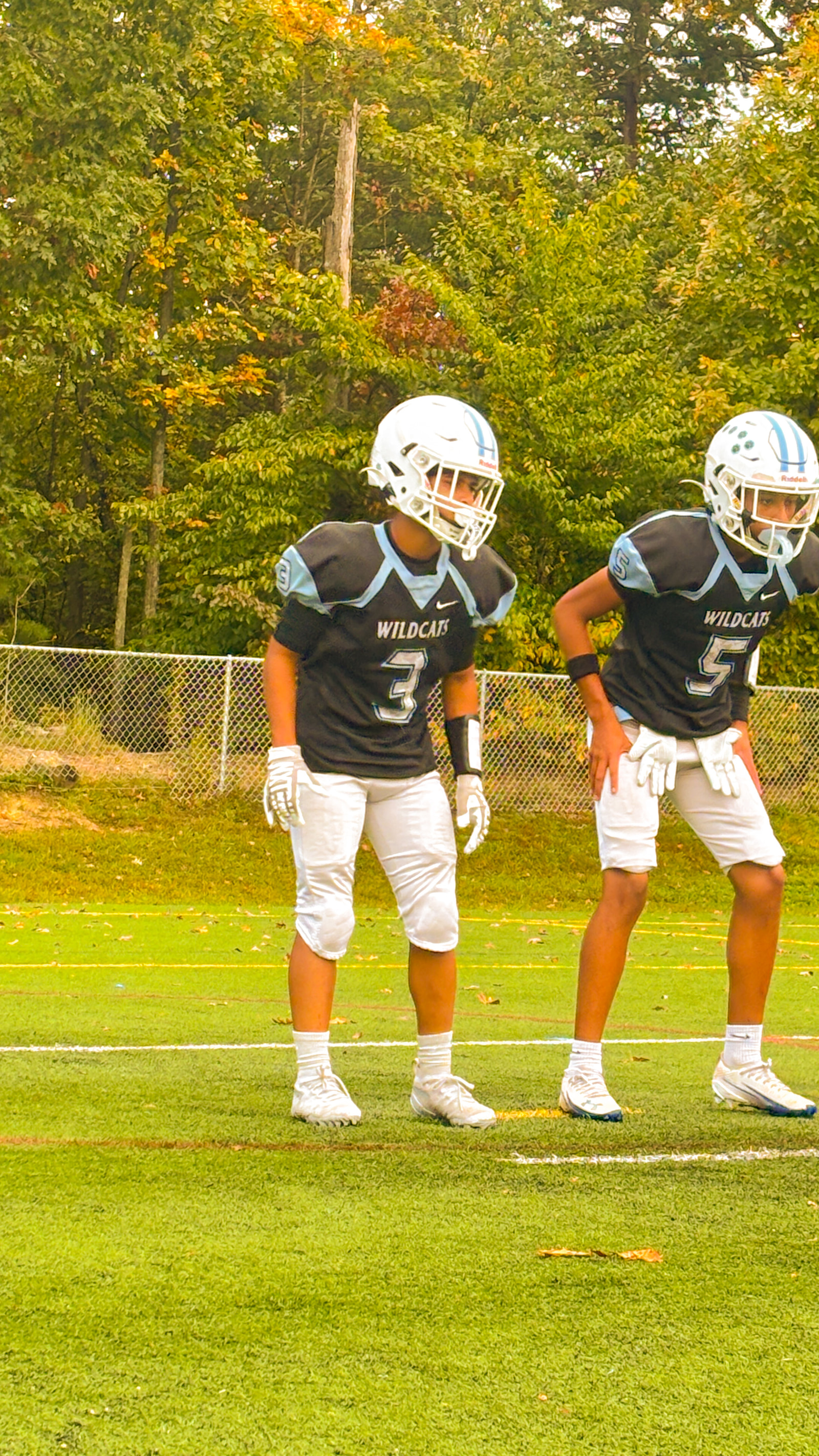 Two young athletes in football gear stand side by side on a grassy field.