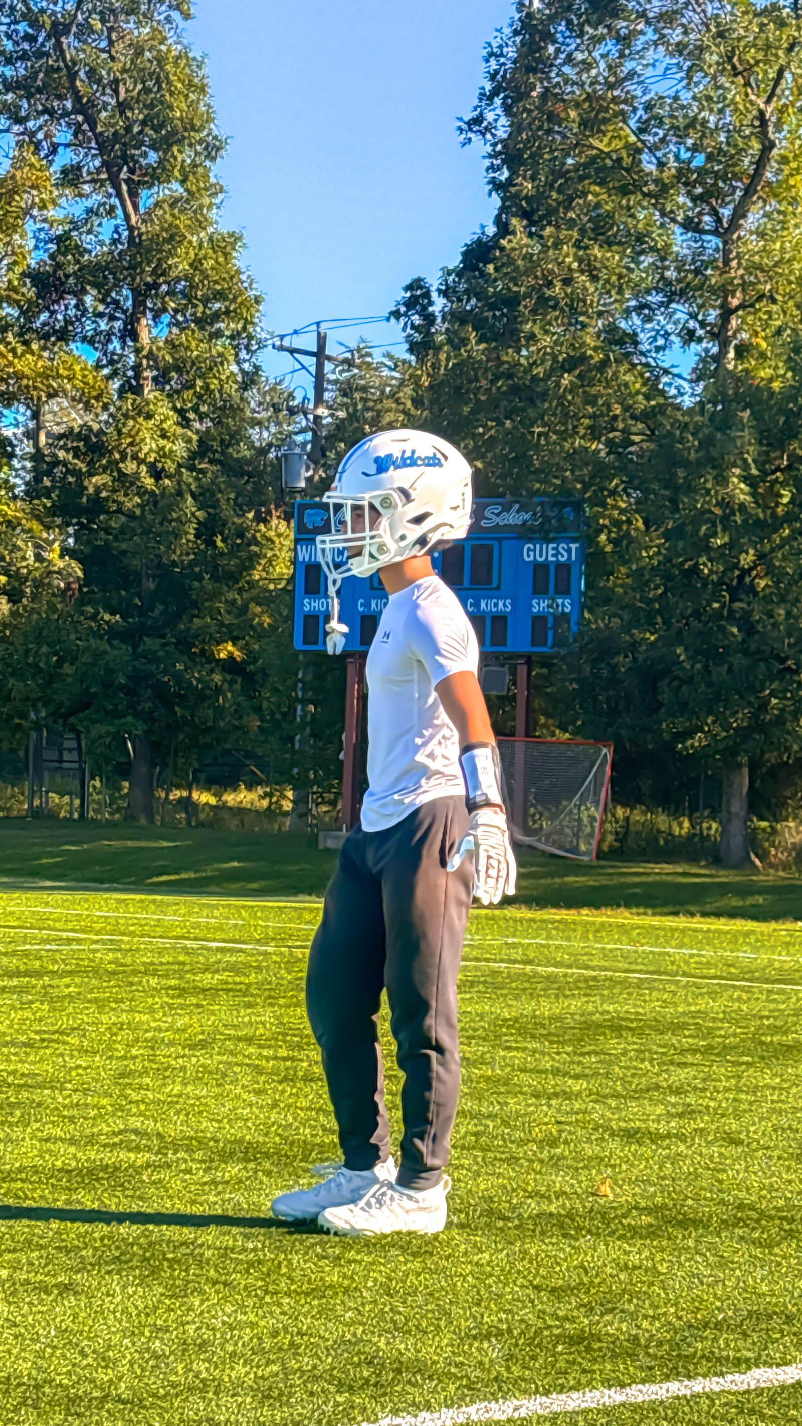 A young athlete stands on a grassy field wearing a white helmet, a light-colored shirt, dark pants, and athletic shoes, with a scoreboard visible in the background.