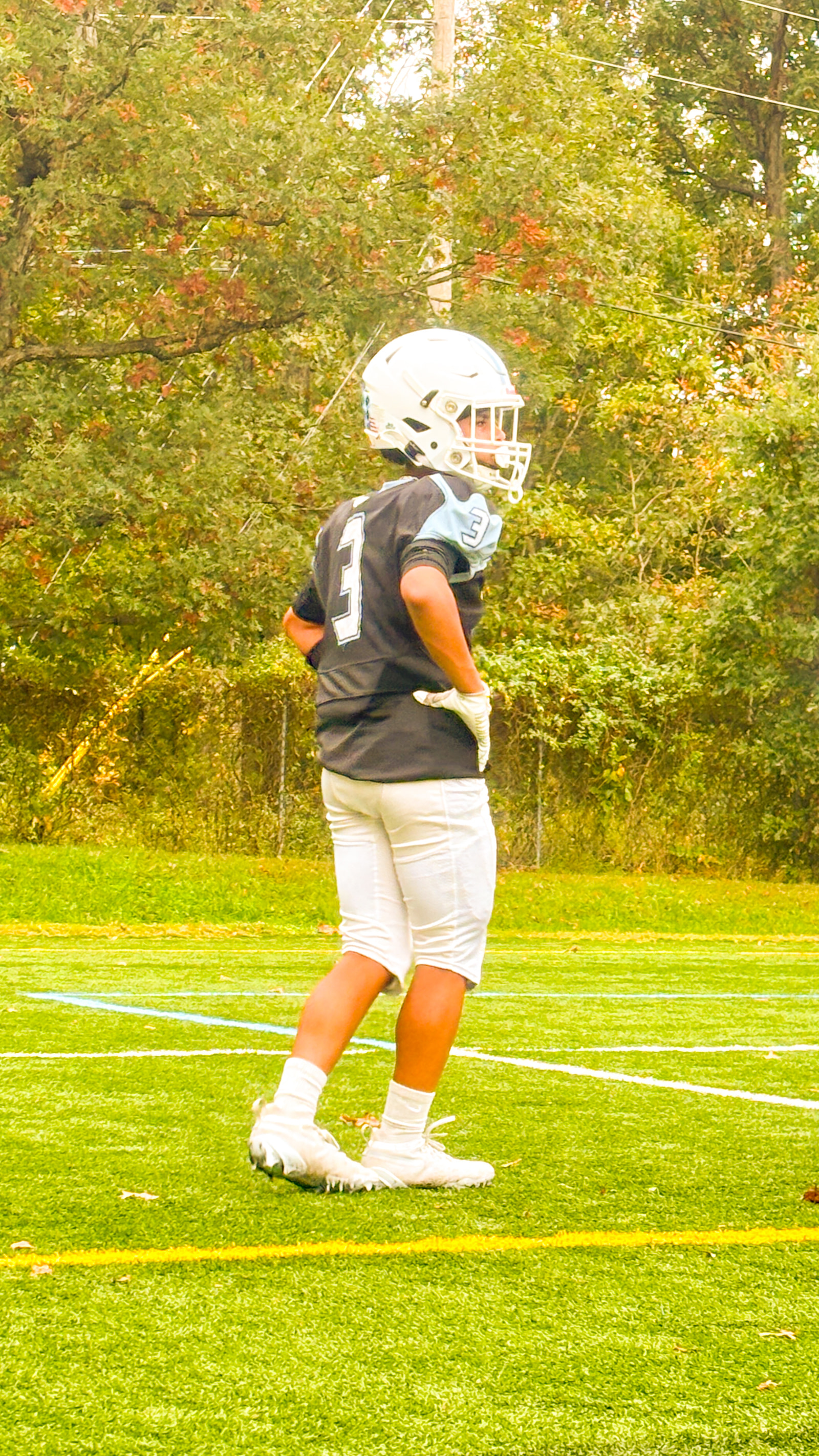 A young athlete in a football uniform stands on a grassy field, facing away from the camera.