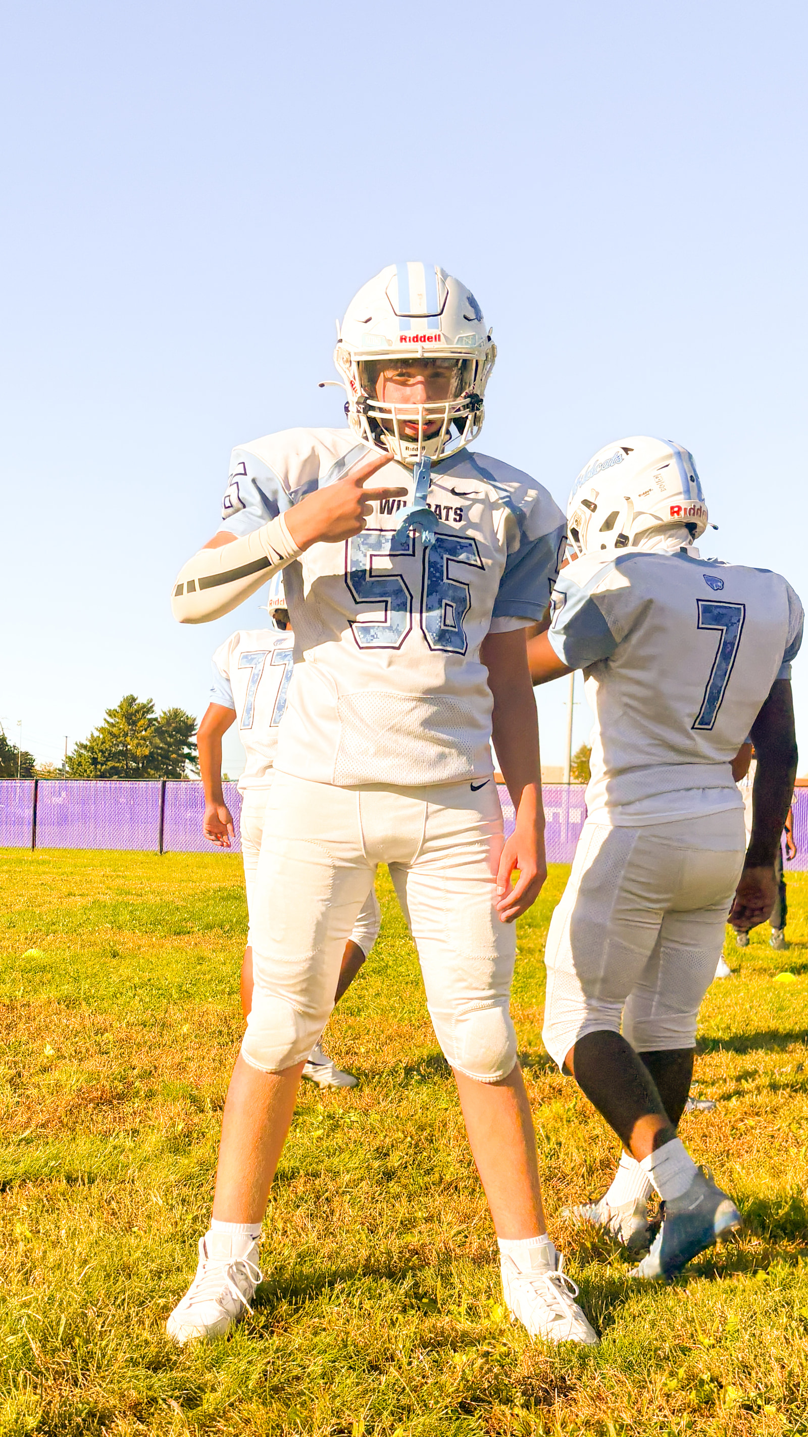 Three teenage athletes in white football uniforms stand on a grassy field, one gesturing with his hand while the others look in different directions.
