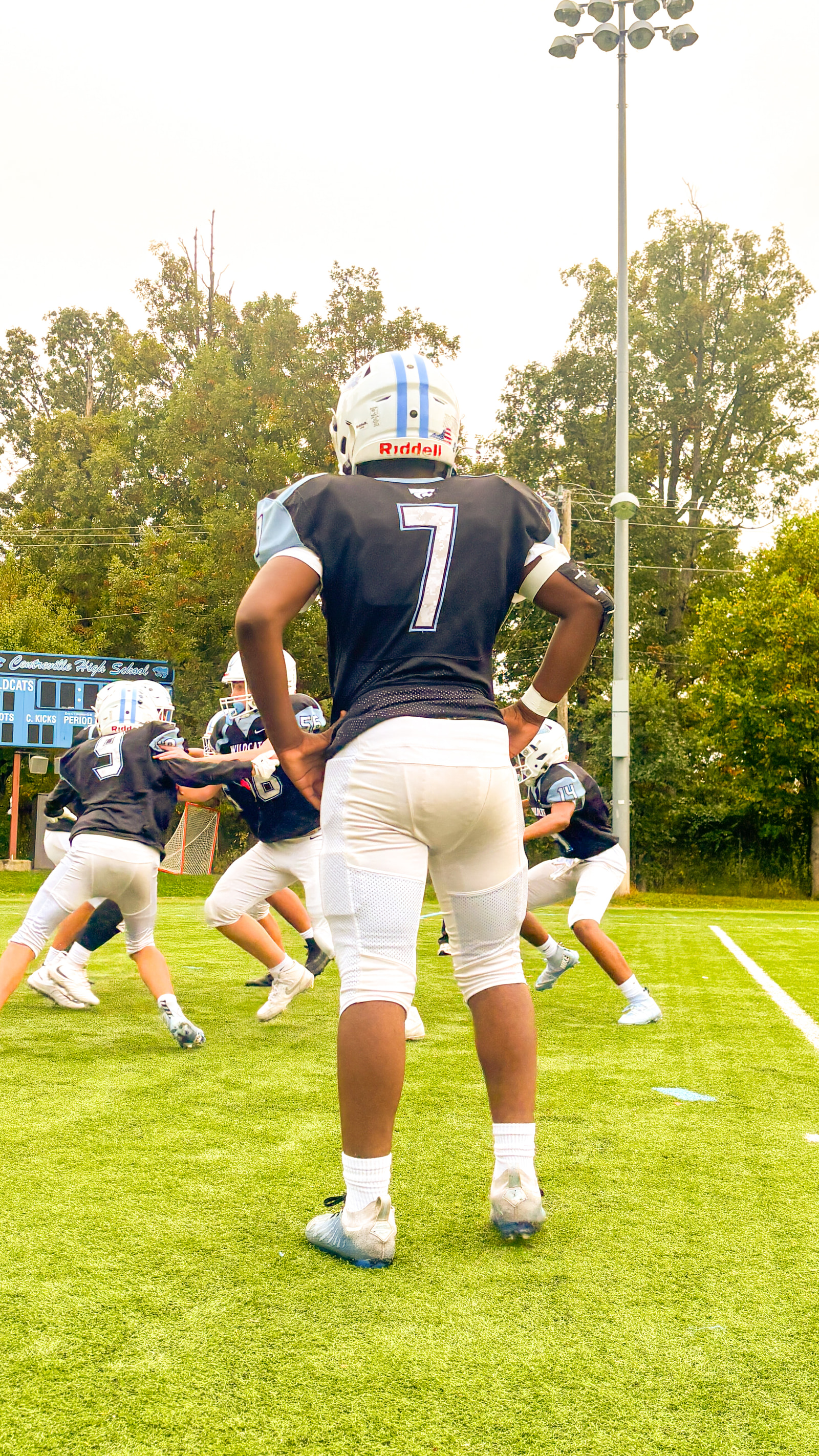 Teenage football players in practice gear are on a field, with one player in the foreground facing away from the camera.