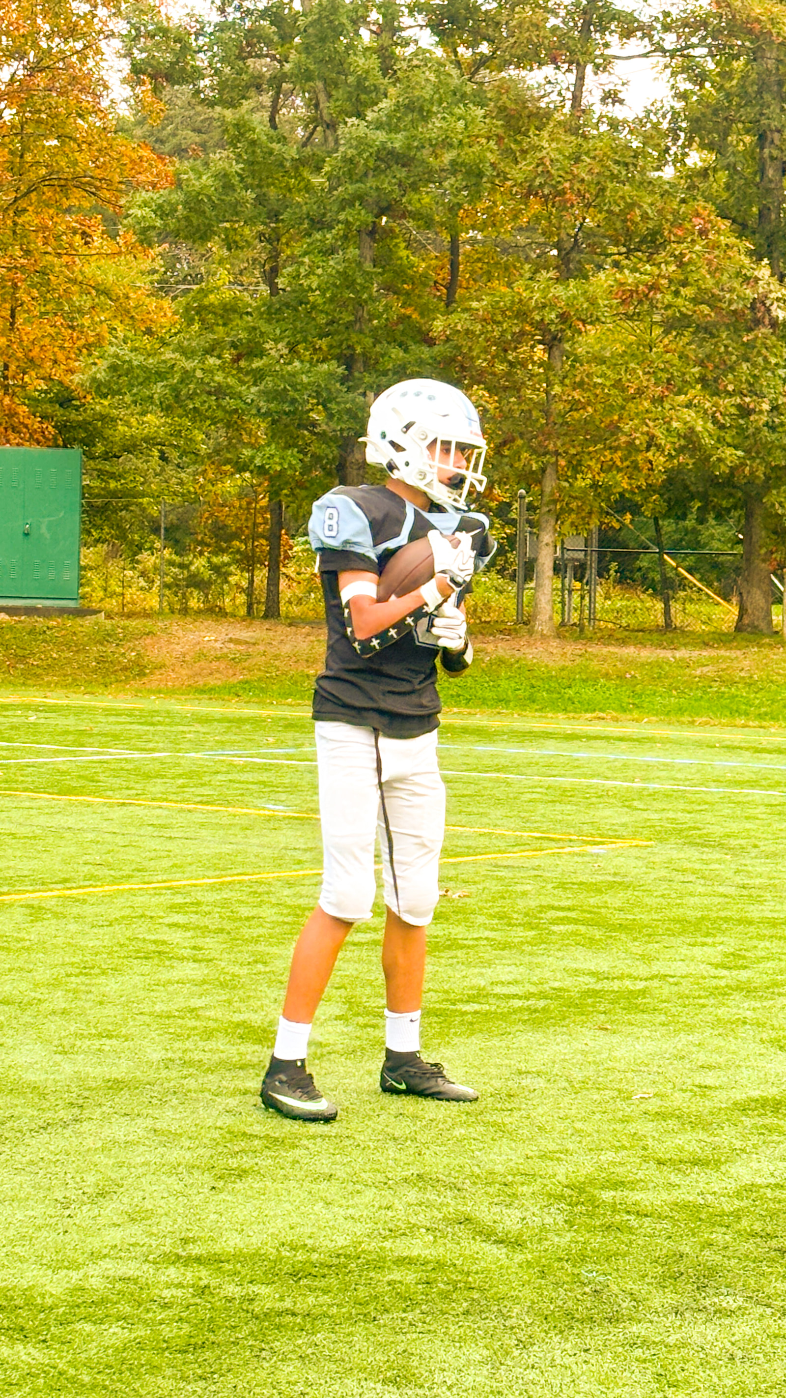 A young athlete in a football uniform stands on a grassy field, holding a football and wearing a helmet.