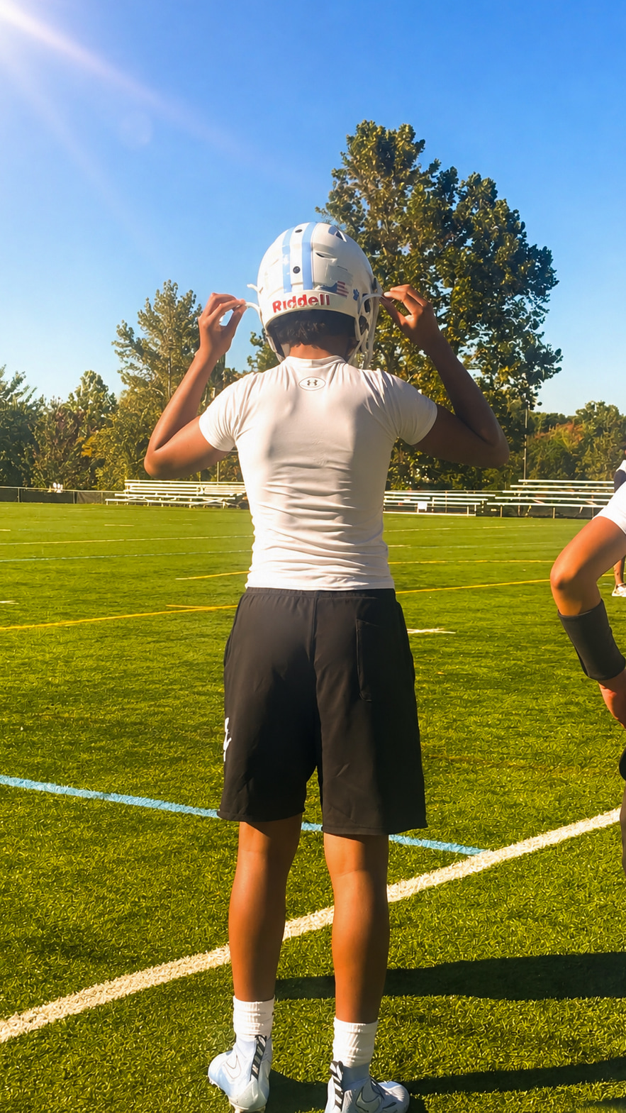 A young athlete adjusts a helmet on a football field, facing away from the camera.