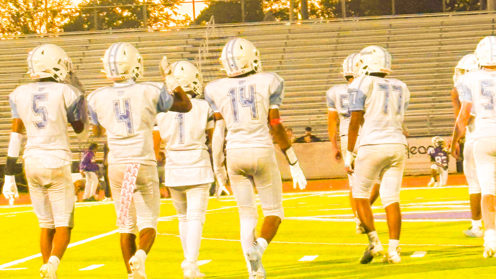 Seven athletes in white jerseys and helmets stand together on a football field, facing away from the camera.