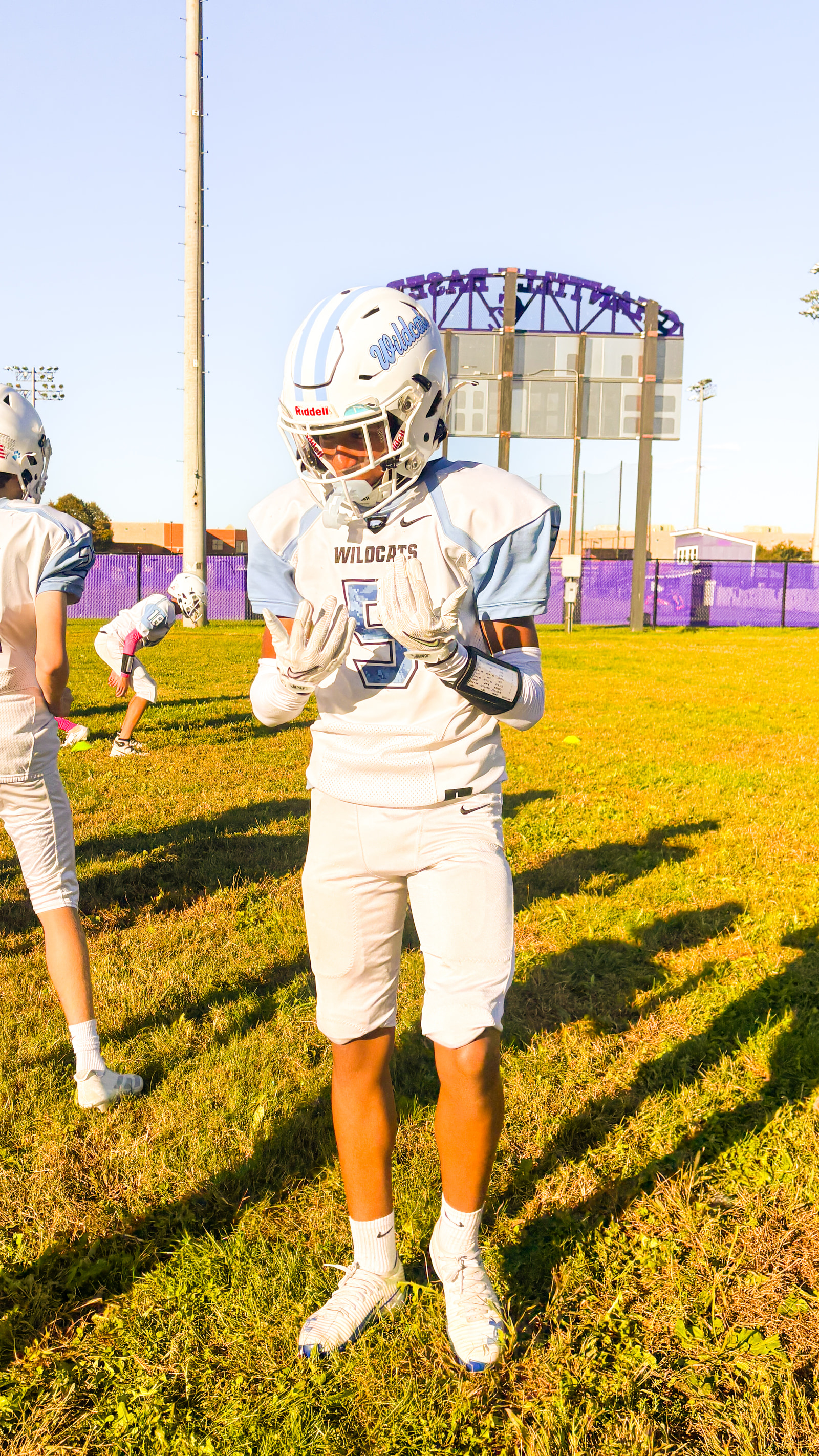 A young athlete in a white and blue football uniform stands on a grassy field, wearing a helmet and gloves, with teammates practicing in the background.