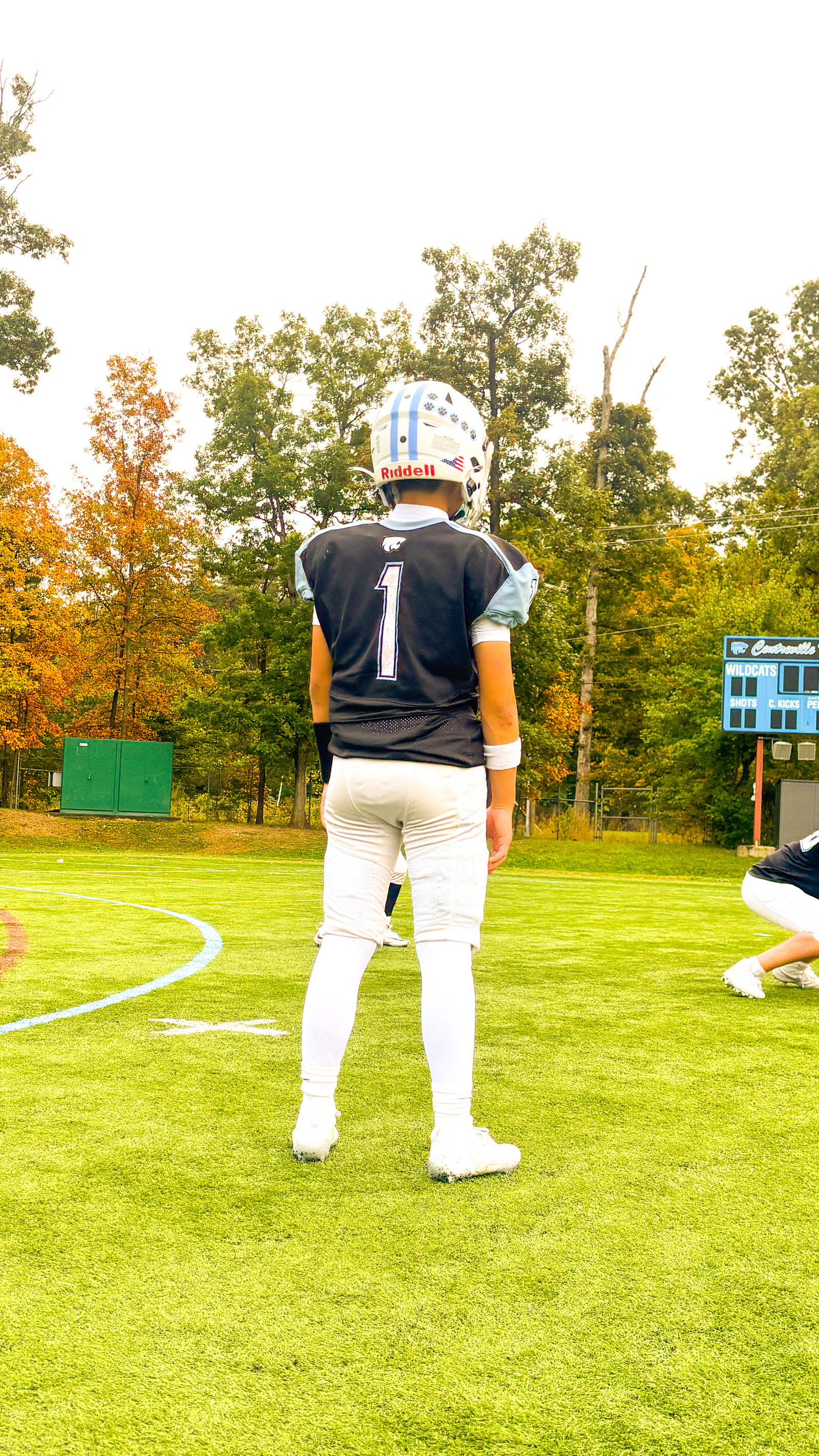 A young athlete stands on a grassy field, facing away from the camera, wearing a dark jersey with the number 1 and a white helmet.