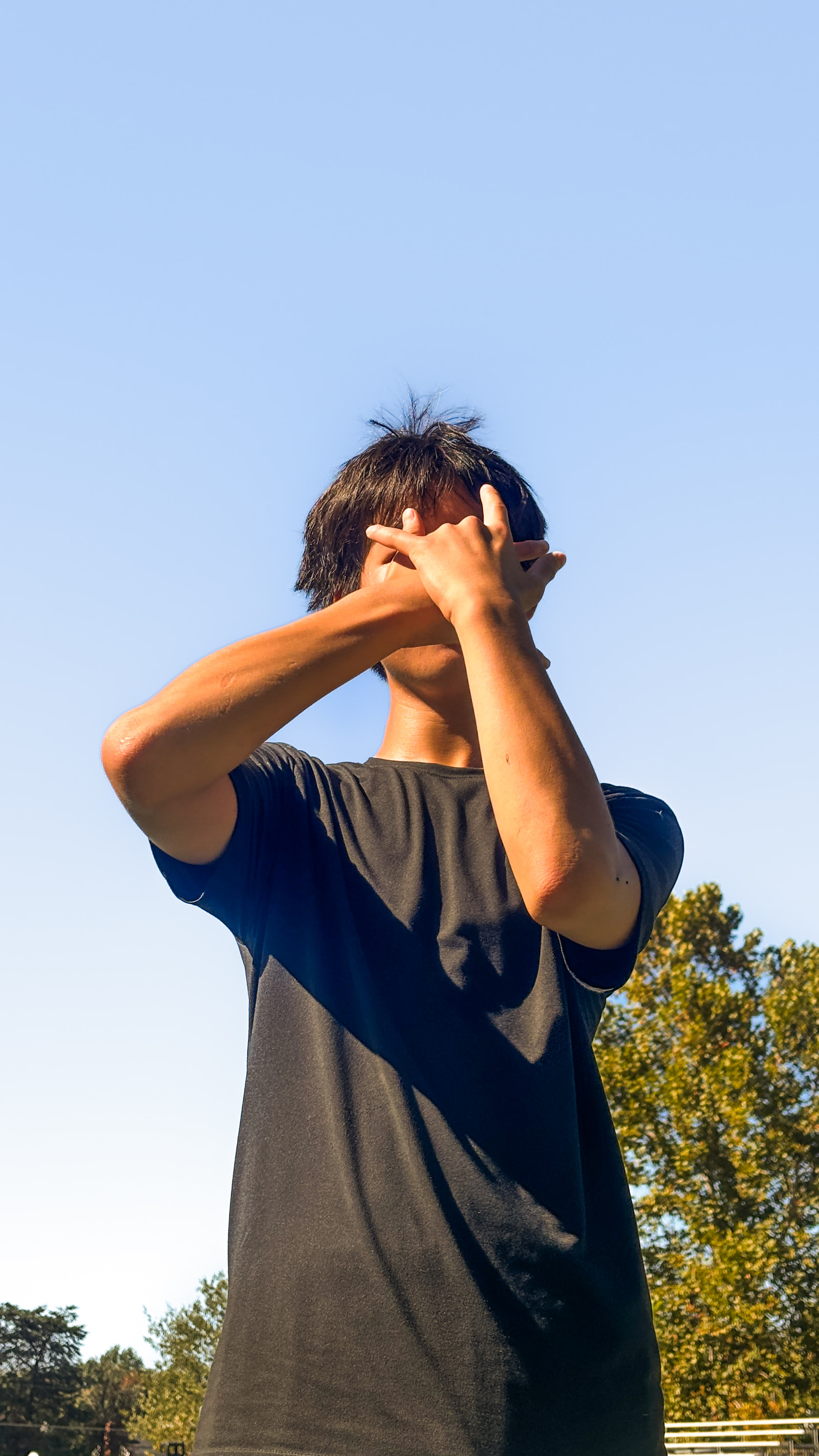 A young man stands with his back to the camera, his hands raised to his head, wearing a dark t-shirt, against a clear blue sky with trees in the background.