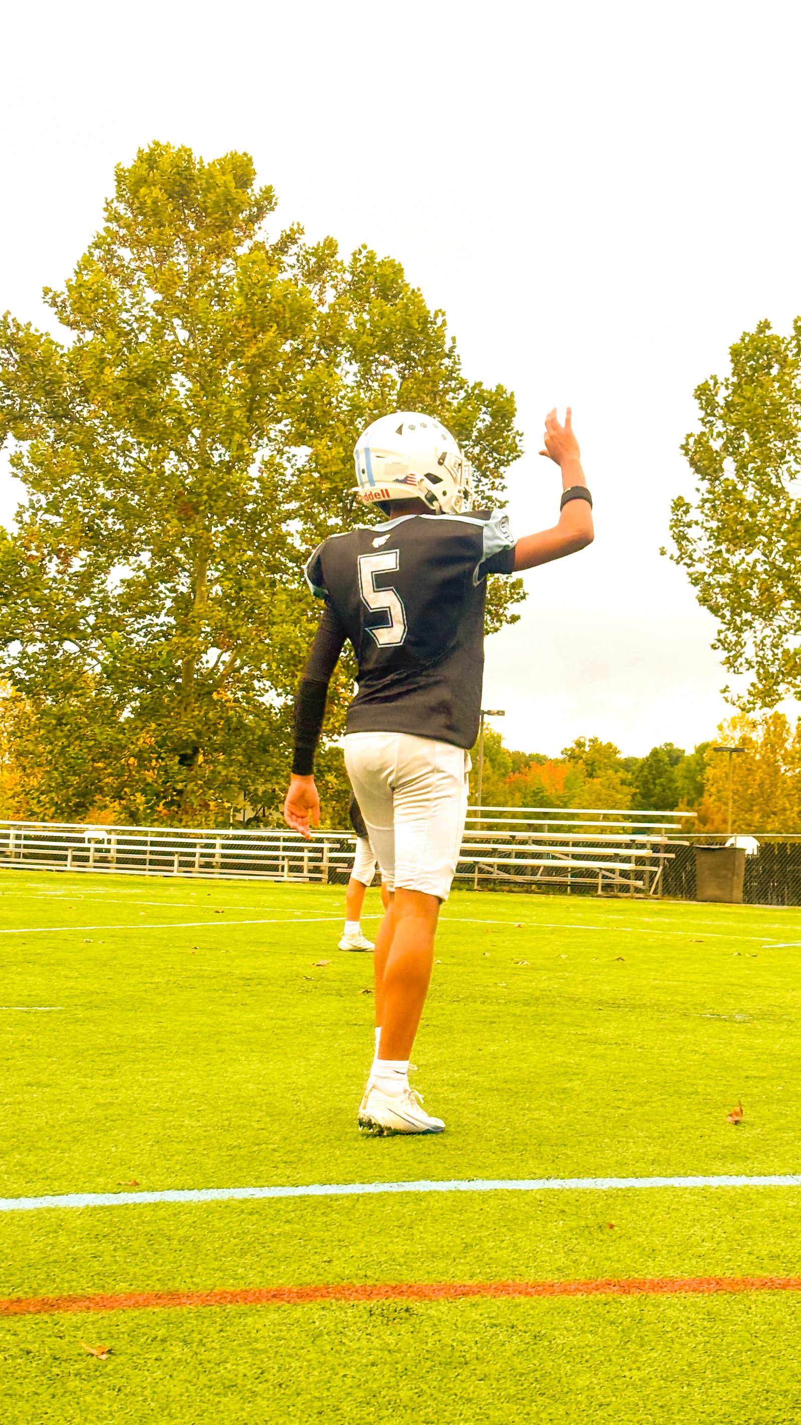 A young athlete in a black jersey with the number 5 stands on a grassy field, raising one arm while wearing a white helmet and white shorts.