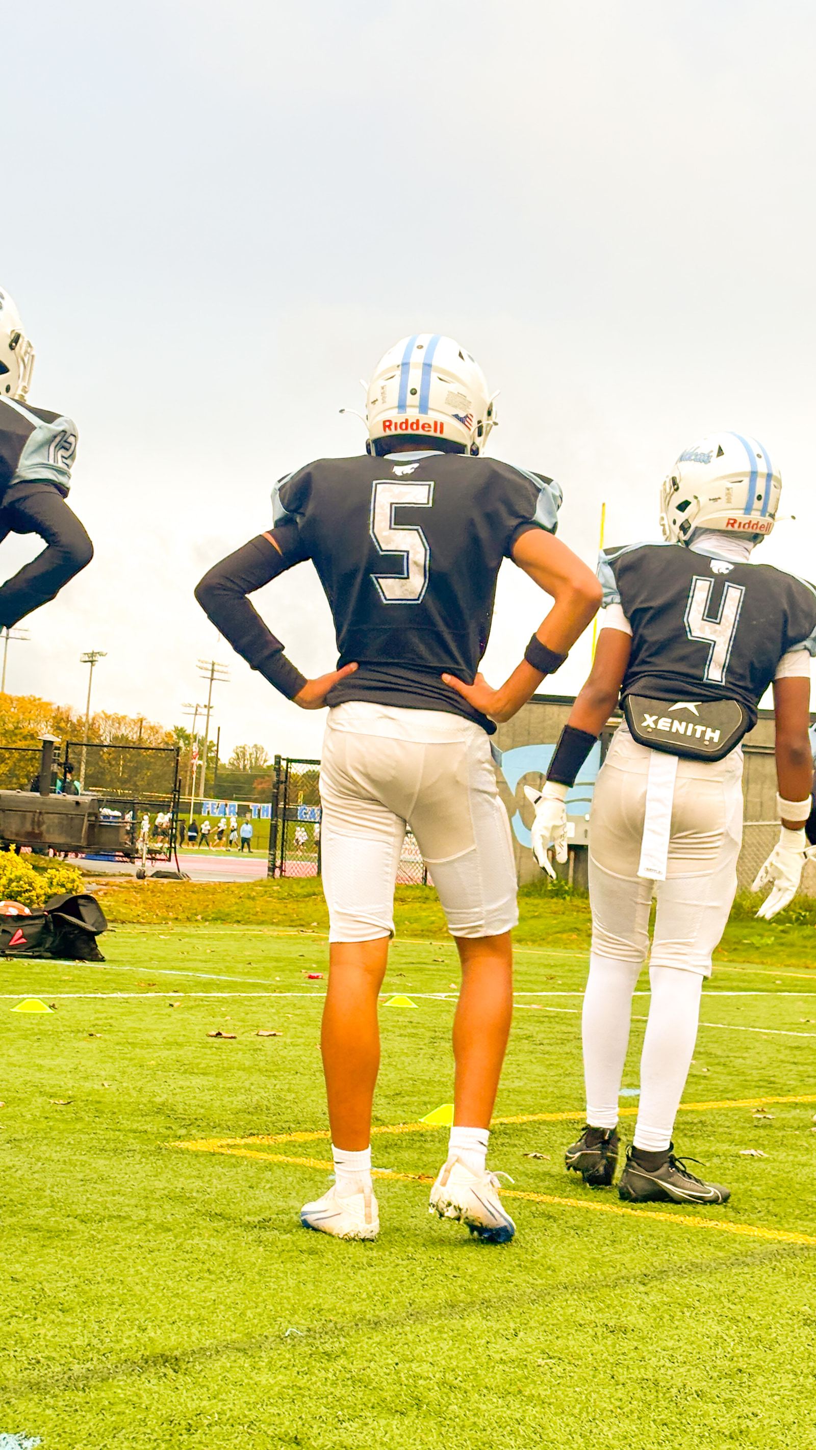 Three football players in dark jerseys and white pants stand on a field, facing away from the camera.