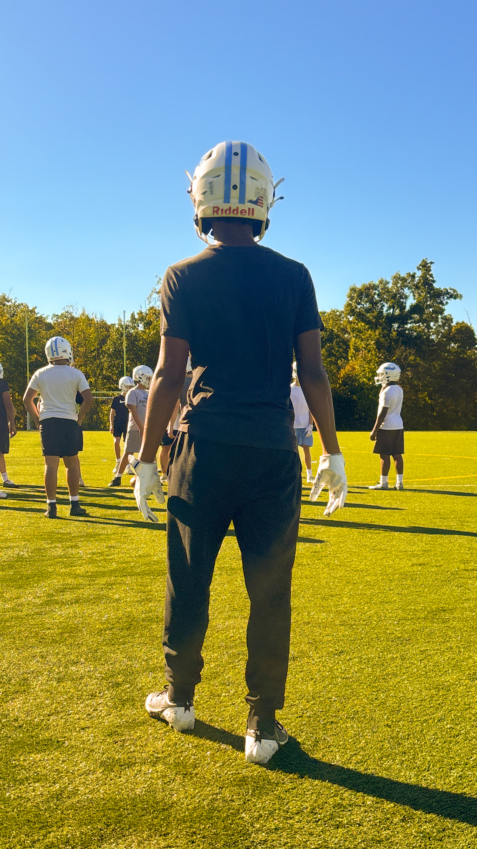 A group of athletes in helmets and gloves stand on a grassy field, facing away from the camera.
