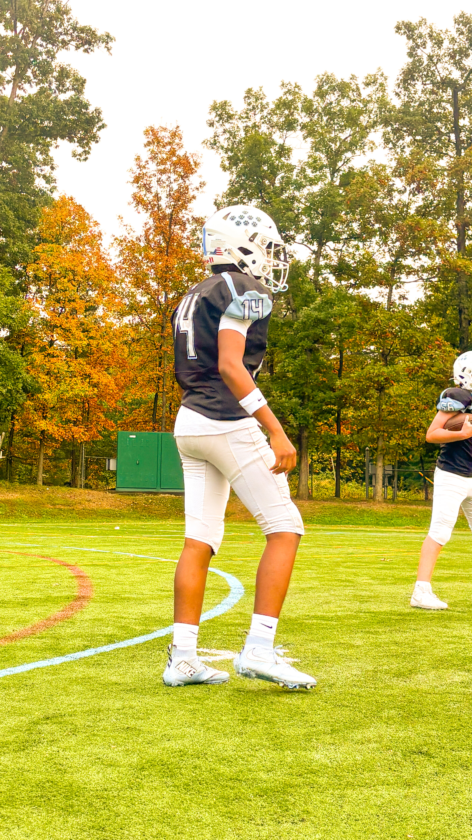 A young athlete in a football uniform stands on a field, facing the camera, with another player visible in the background.