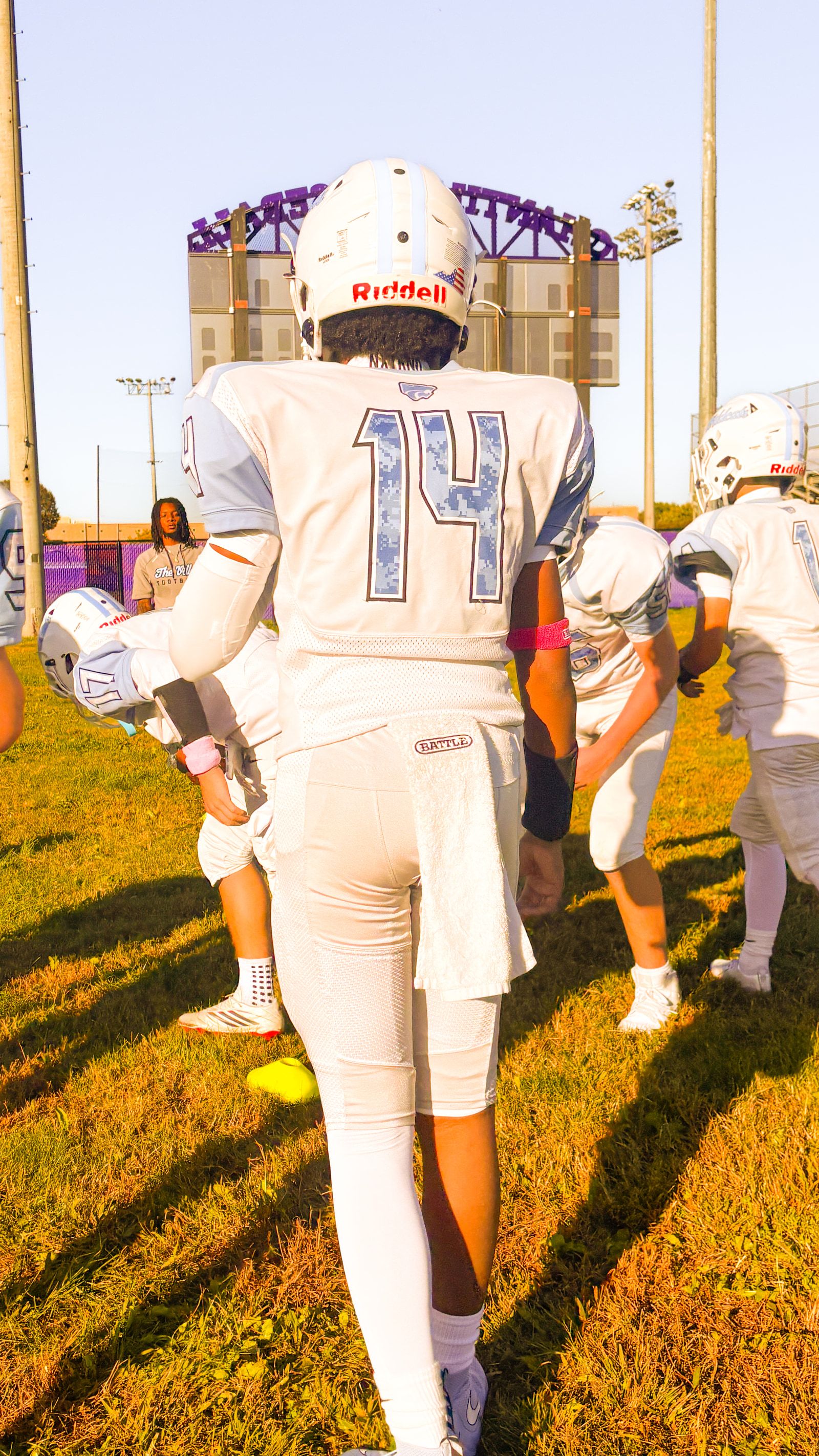 Teenage football players in white jerseys and helmets stand on a grassy field, facing away from the camera.