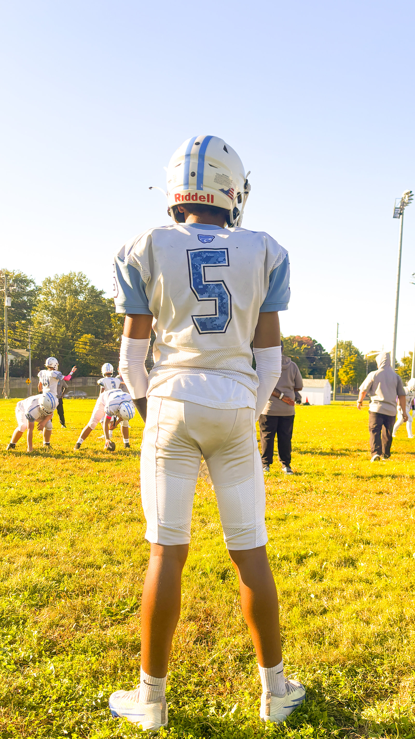 A young athlete in a white jersey with the number 5 stands on a grassy field, facing away from the camera while teammates practice in the background.