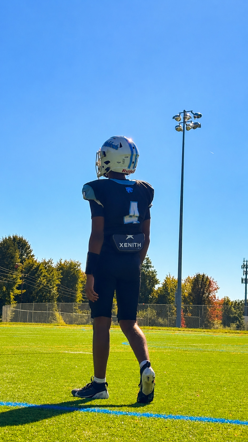 A young athlete stands on a sports field, facing away from the camera, wearing a helmet and a dark jersey with the number 4.