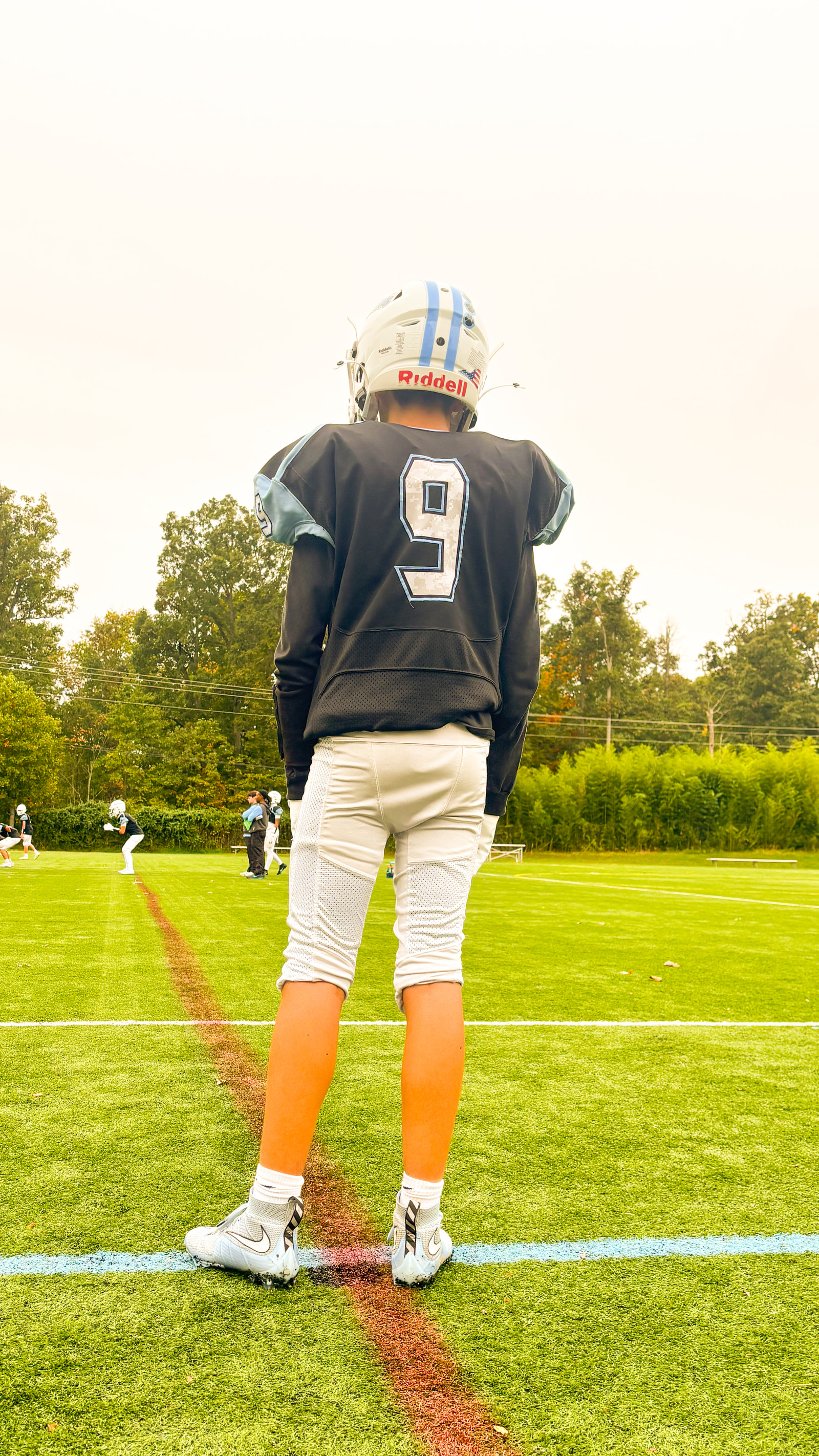 A young athlete in a football uniform stands on a field, facing away from the camera, with teammates practicing in the background.