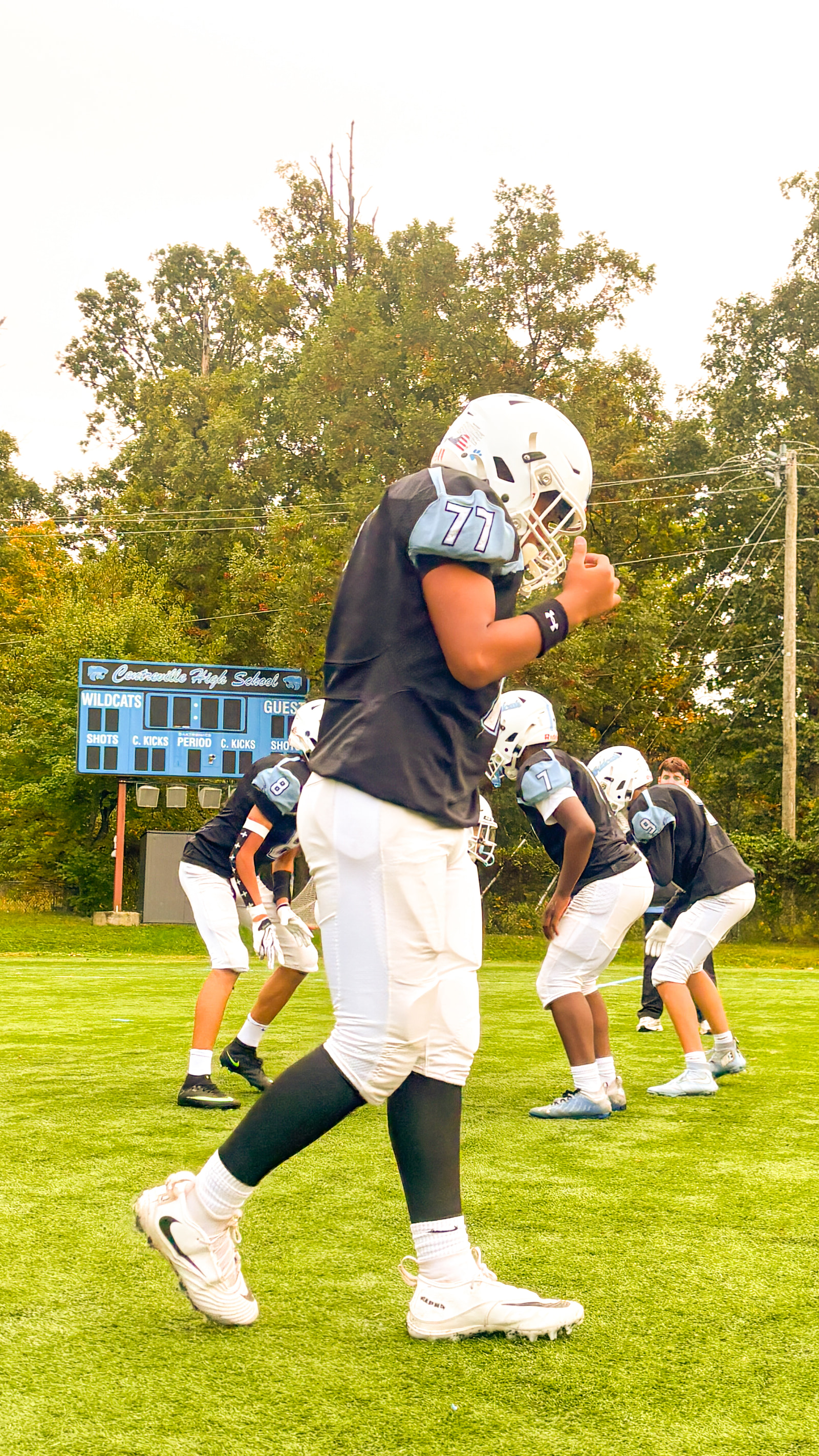 Teenage football players in practice gear stand on a field, with one player in the foreground giving a thumbs-up.