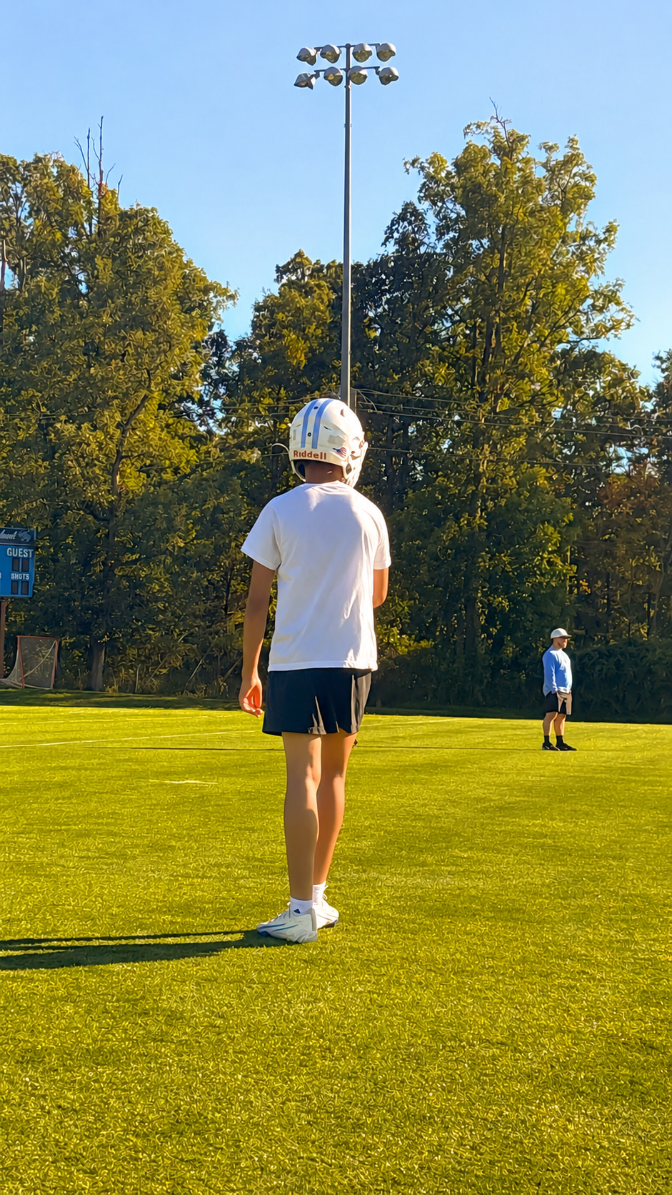 A young person in a helmet stands on a grassy field facing a goalpost, with another person in the distance.