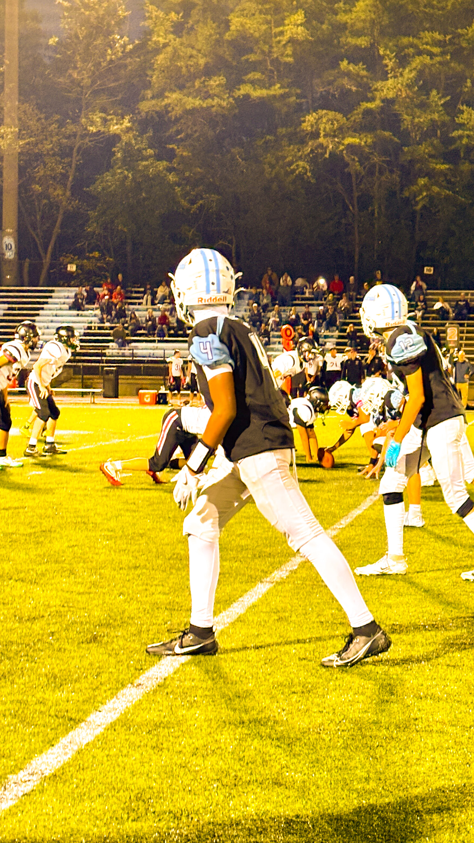 Teenage football players in helmets and uniforms line up on a field during a game at night, with spectators in the stands.