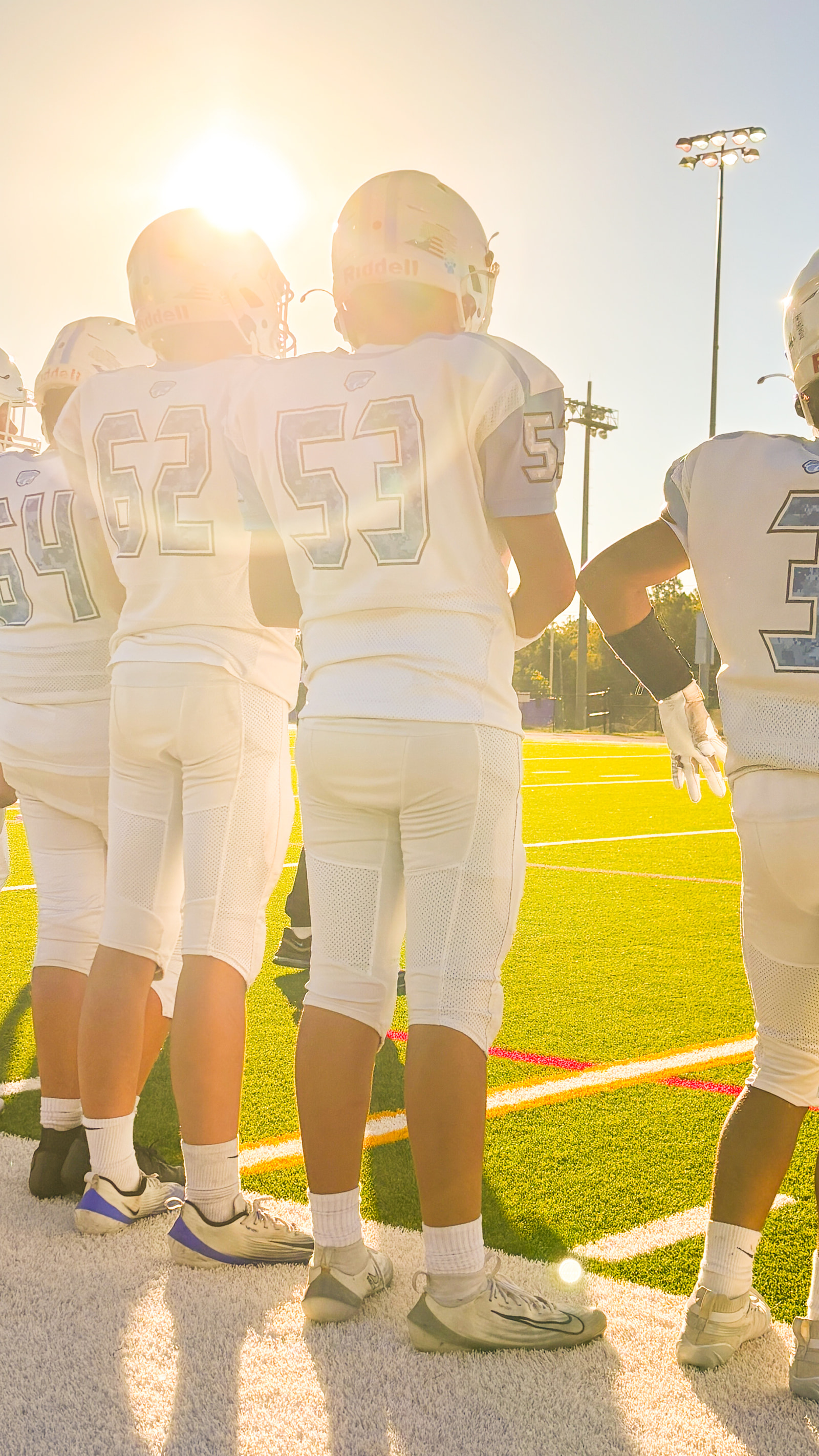 Teenage football players in white jerseys and helmets huddle on a field during sunset.