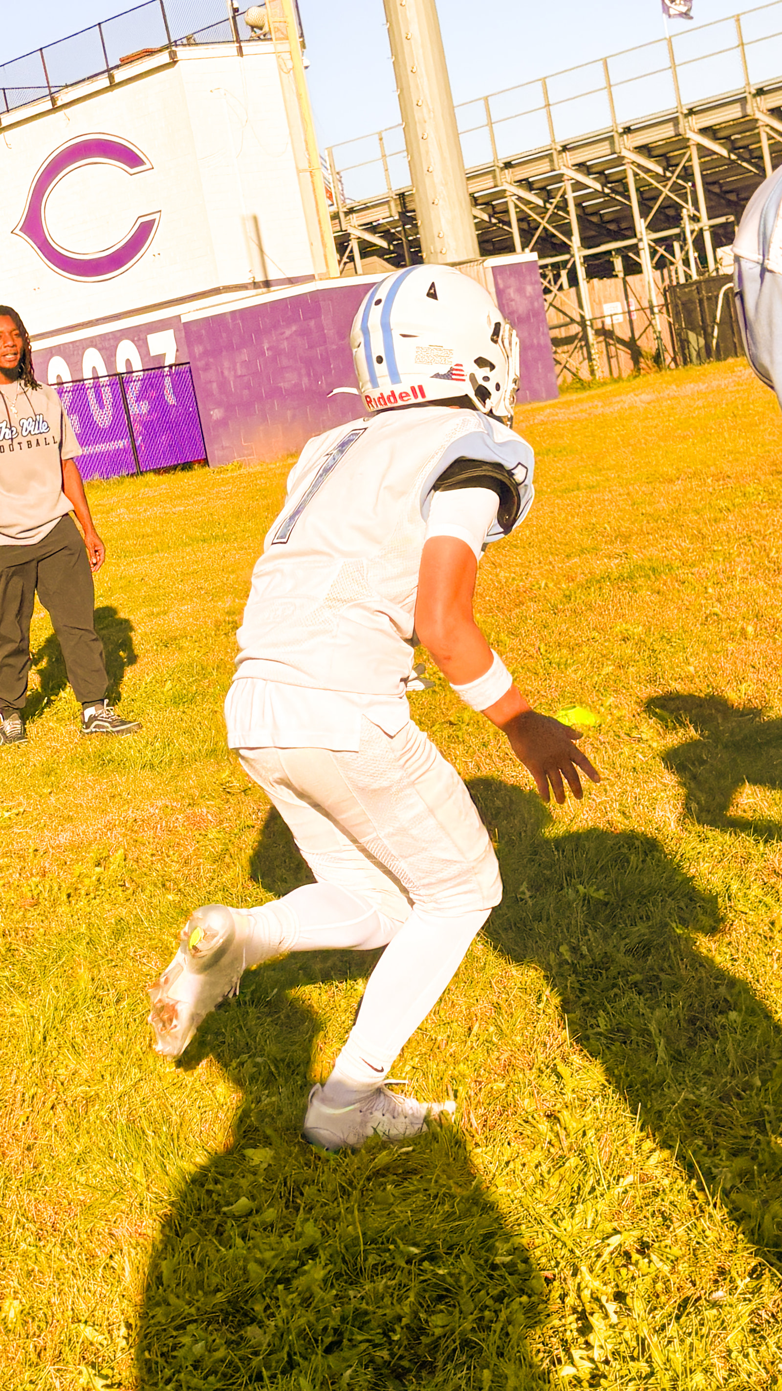 A young athlete in a white uniform and helmet runs with a football on a grassy field, while an adult stands nearby.