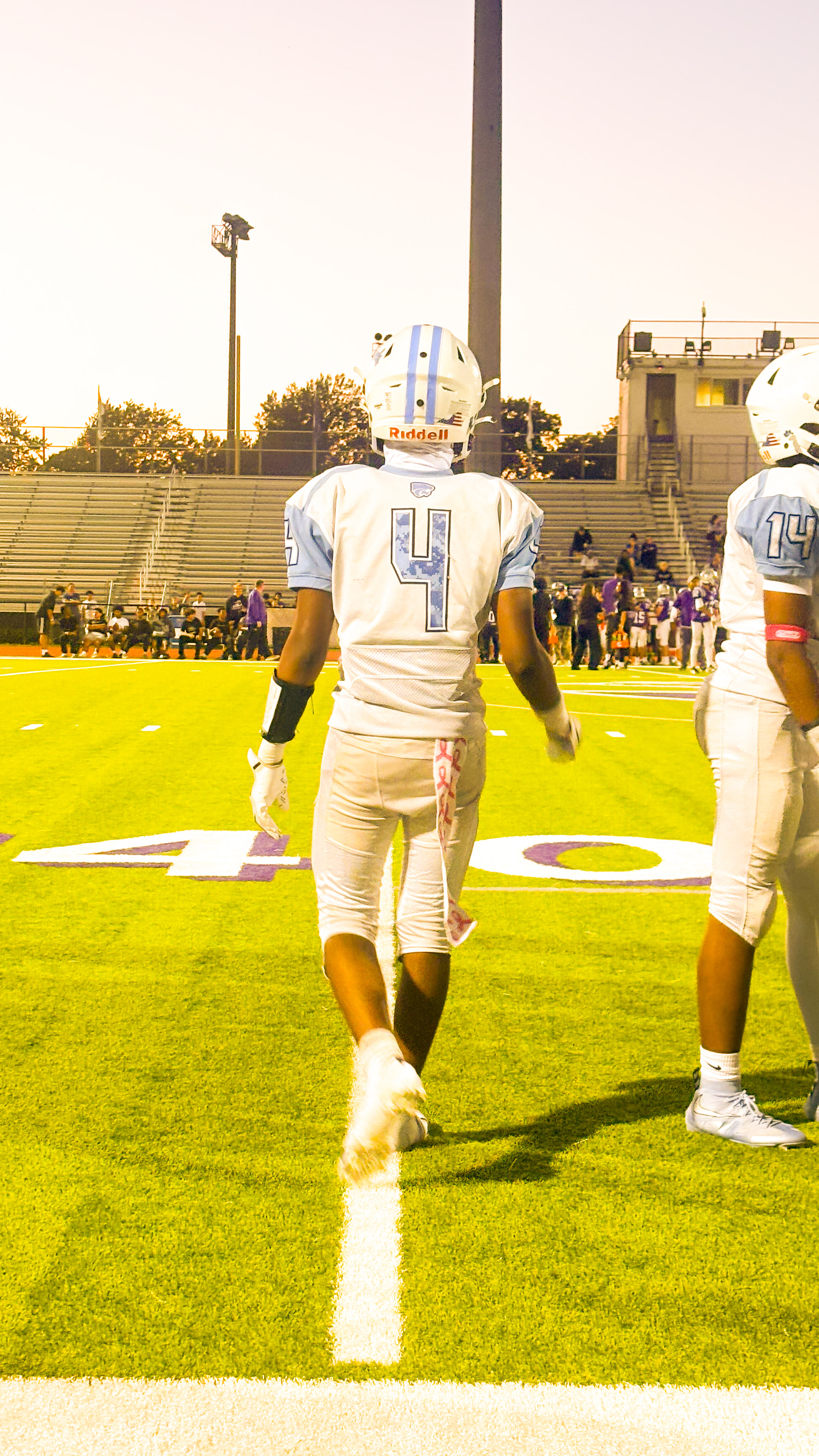 Two athletes in white football uniforms with blue accents stand on a field, facing away from the camera.