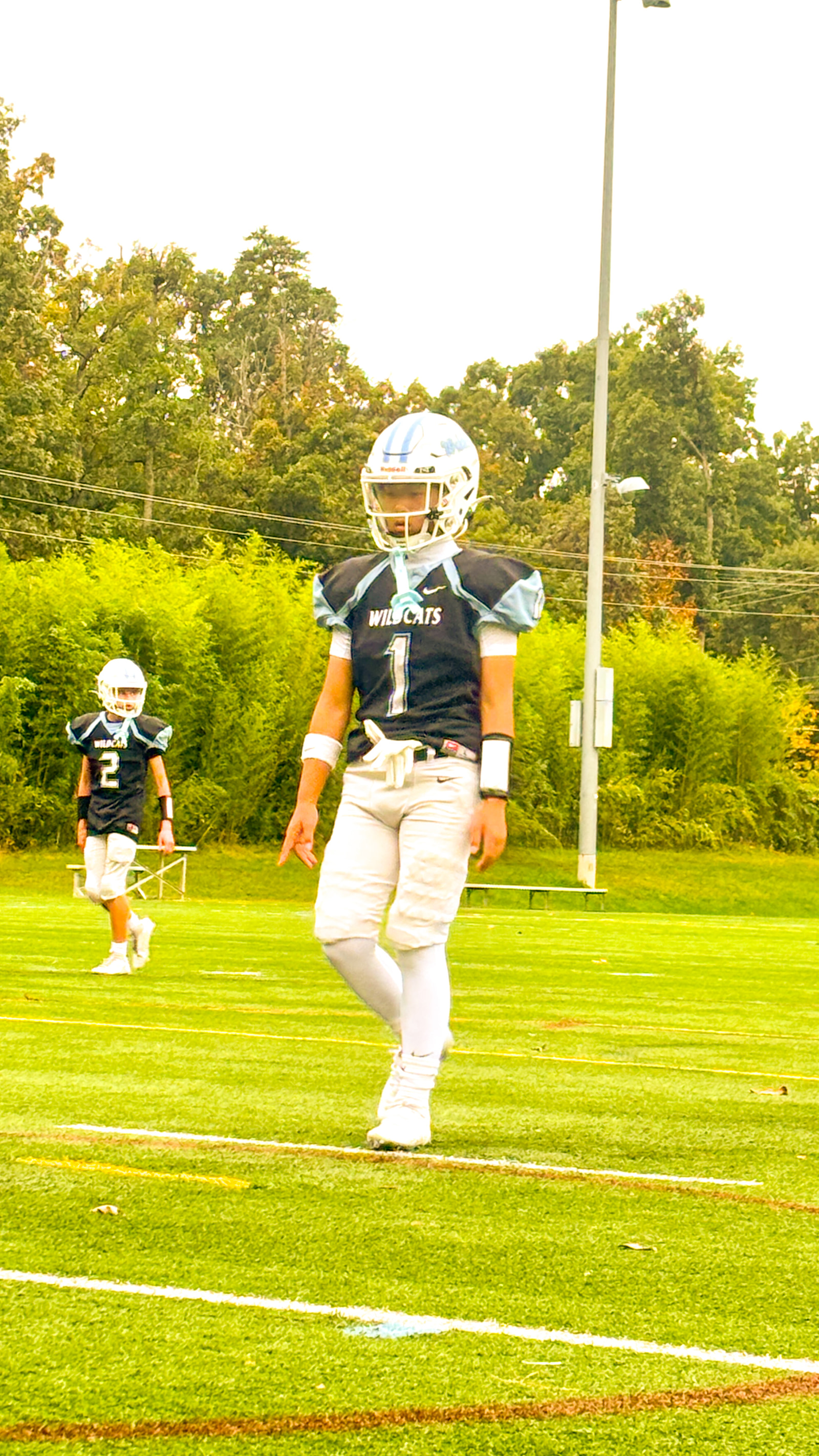 Two young athletes in football gear stand on a field, one in the foreground and the other slightly behind.